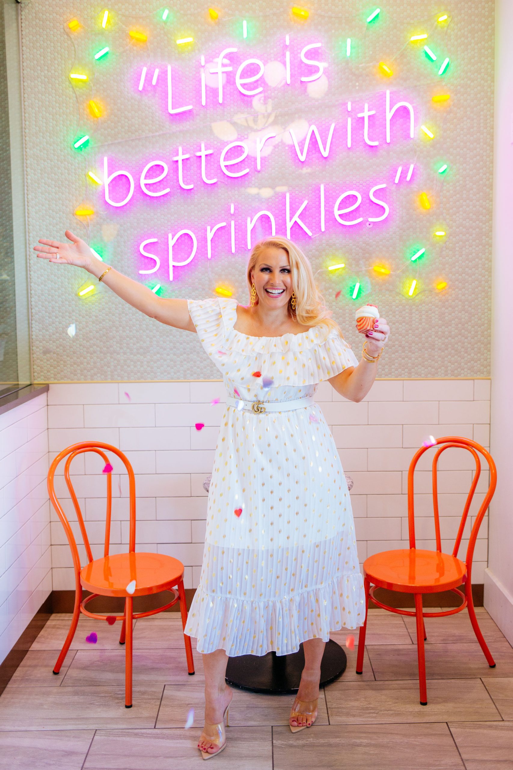 Tampa Fashion blogger Jenn Truman @jtstjtst11 is holding a cupcake and throwing heart confetti as she stands in from of the lit sign that says Life is Better with Sprinkles. She is wearing a white off the shoulder dress with gold dots from Amazon fashion, clear heels and Gold Lisi Lerch earrings.