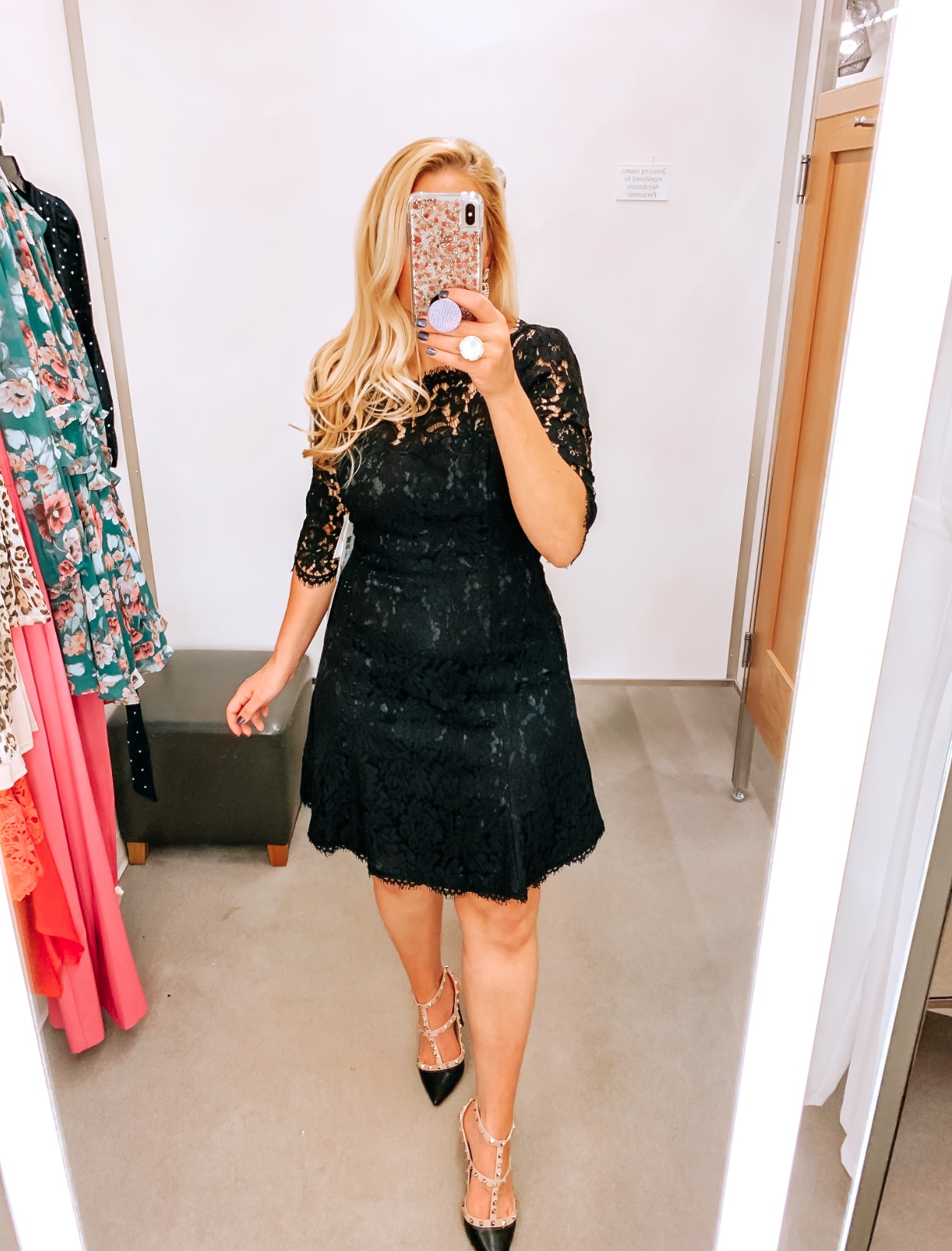 Tampa Blogger Jenn Truman providing a mirror selfie inside Nordstrom at Tampa International Plaza. She is wearing an Eliza J black Lace Fit and flare cocktail dress from Nordstrom and Valentino dupe black studded heels from Amazon. She is holding her phone for a mirror selfie try on session.