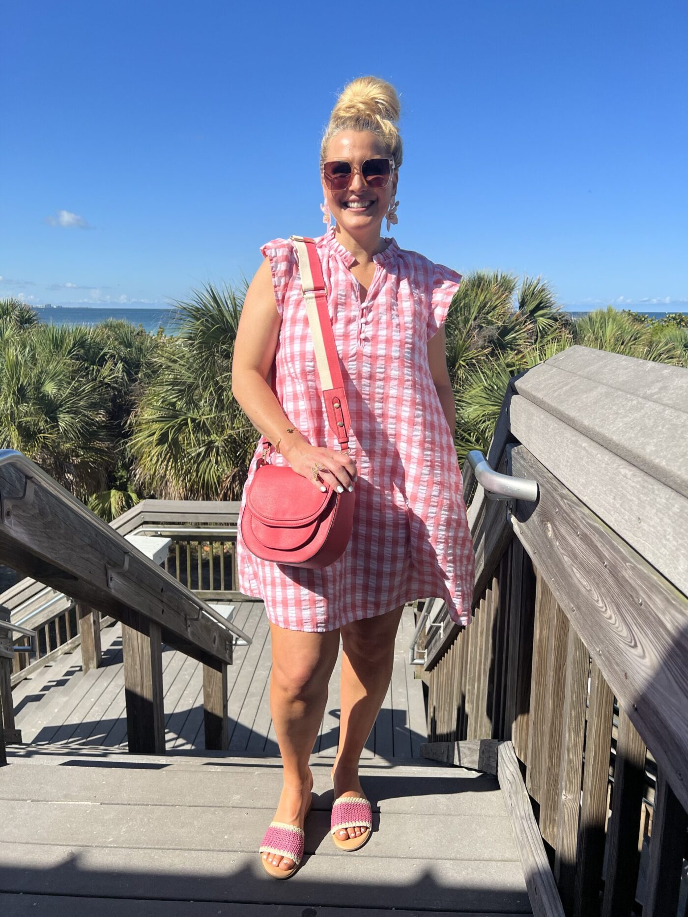 Jenn Truman, Fashion Blogger @JTSTJTST11 is standing on a stair case in Dunedin, FL wearing a pink and white gingham flutter sleeve mini dress while smiling.