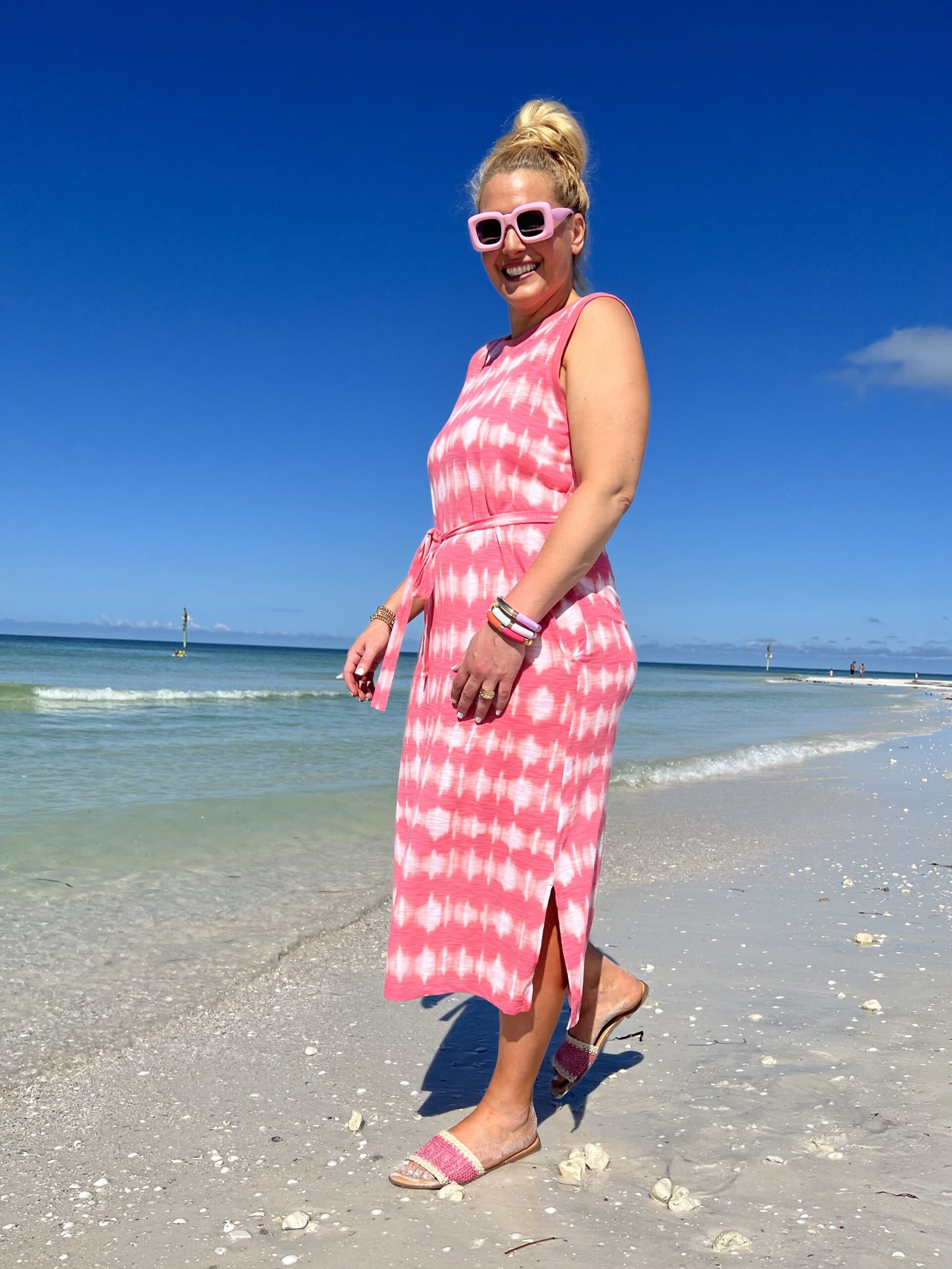 Jenn Truman, Style Blogger @JTSTJTST11 is standing on the beach in Dunedin, FL wearing a sleeveless Pink and white print midi dress and pink rectangle sunglasses. She's near the ocean while smiling.